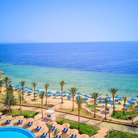 Pickalbatros Royal Grand Sharm - Adults Friendly 16 Years Plus Екстериор снимка The photo displays a beautiful coastal scene featuring a clear blue sea that transitions into green tones near the shore. In the foreground, there are sandy beaches lined with palm trees and a variety of beach umbrellas. There is also a swimming pool