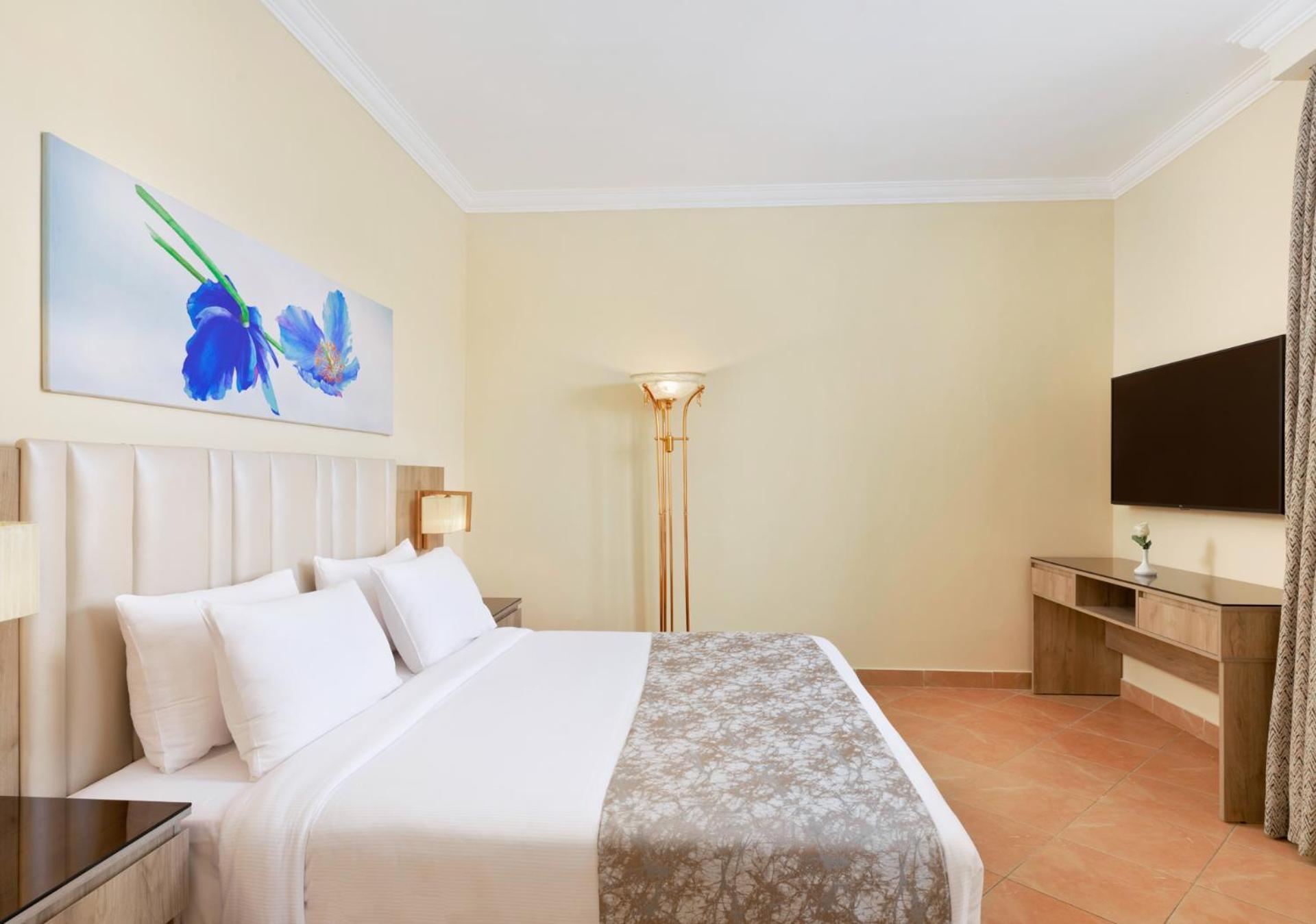 Pickalbatros Royal Grand Sharm - Adults Friendly 16 Years Plus Екстериор снимка The photo shows a modern hotel room featuring a large bed with white linens and a decorative gray throw. On one side of the room, there is a piece of artwork depicting blue flowers. A floor lamp provides additional lighting, and across from the bed i