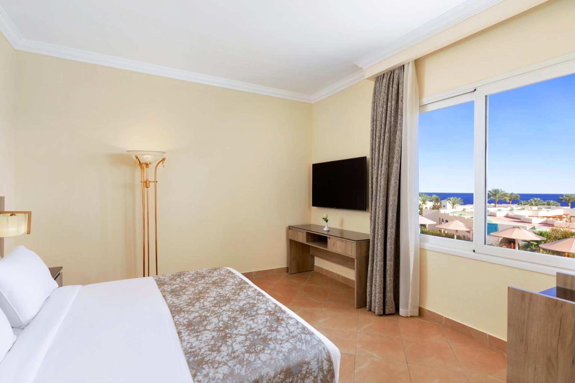 Pickalbatros Royal Grand Sharm - Adults Friendly 16 Years Plus Екстериор снимка The photo shows a hotel room with a modern and minimalistic design. There is a comfortable bed with white linens and a decorative gray throw at the foot. A large window offers a view of the outdoors, possibly with a glimpse of the sea or beach. A fla