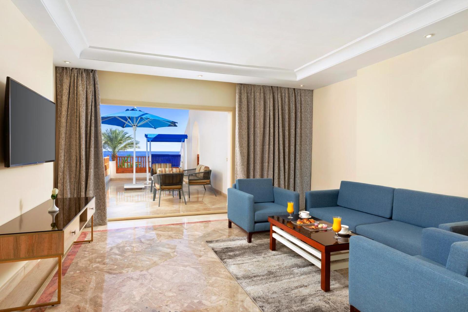 Pickalbatros Royal Grand Sharm - Adults Friendly 16 Years Plus Екстериор снимка The photo shows a spacious living area with modern decor. There are two blue sofas arranged around a coffee table that holds some decorative items and candles. A flat-screen TV is mounted on the wall across from the seating area. To the left, there a