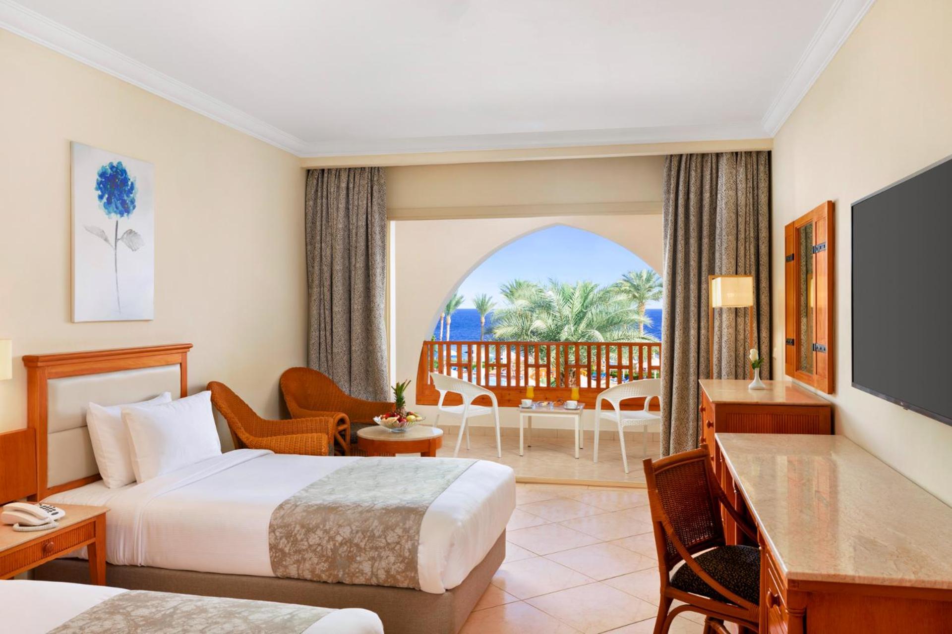 Pickalbatros Royal Grand Sharm - Adults Friendly 16 Years Plus Екстериор снимка The photo shows a hotel room featuring two beds with light bedding. There is a wooden desk and chair, and a comfortable chair is placed nearby. The room has large windows that open to a balcony with a view of palm trees and a blue sky. The decor is r