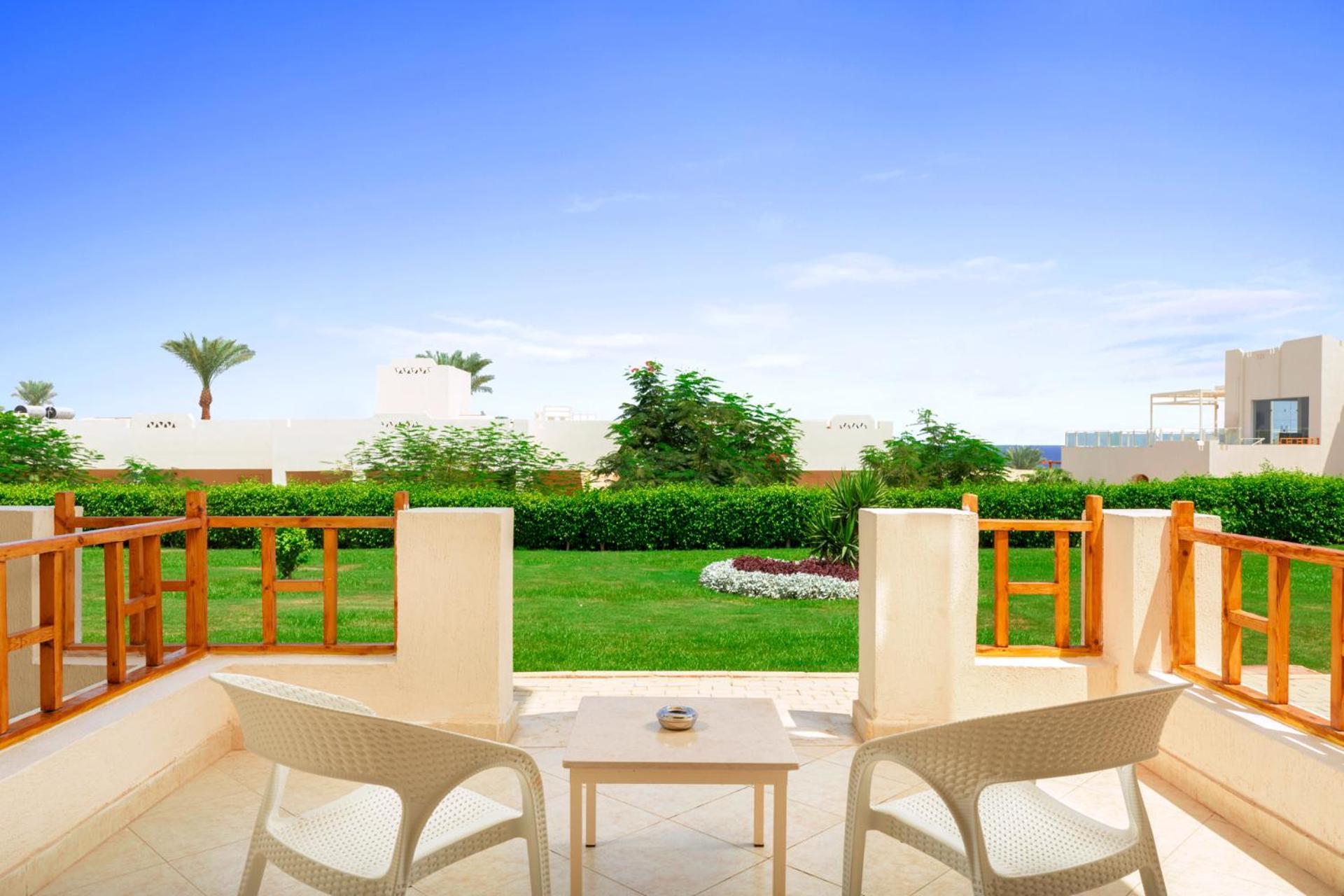 Pickalbatros Royal Grand Sharm - Adults Friendly 16 Years Plus Екстериор снимка The photo depicts a serene outdoor balcony or patio area. It features two white chairs and a small table, set against a backdrop of lush green grass and garden plants. In the distance, there are some palm trees and clear blue skies, suggesting a warm