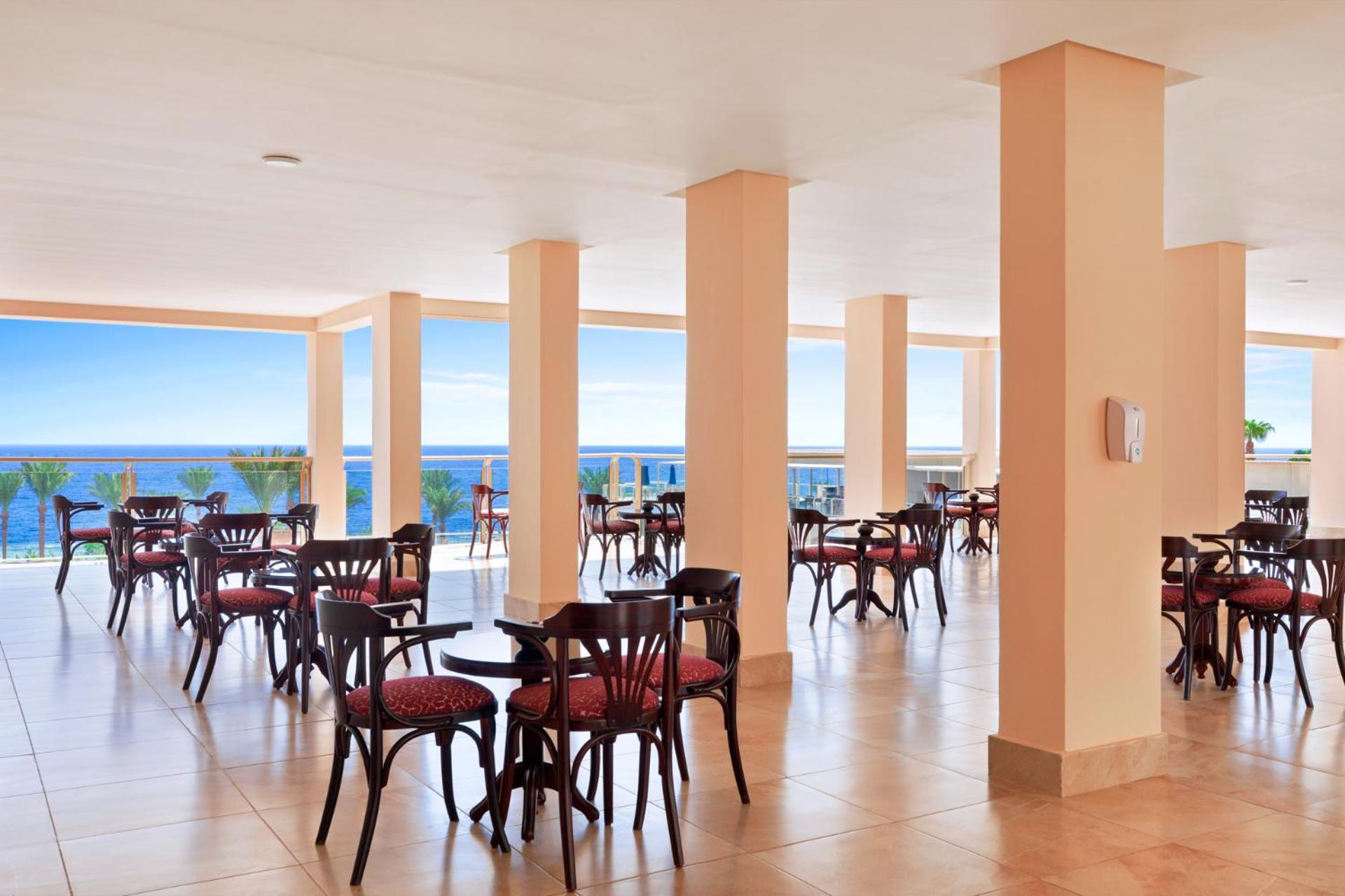 Pickalbatros Royal Grand Sharm - Adults Friendly 16 Years Plus Екстериор снимка The image shows a dining area or café with several round wooden tables and chairs, featuring red upholstery. The space is well-lit, with large windows that allow a view of the ocean in the background, contributing to a bright and open atmosphere. The