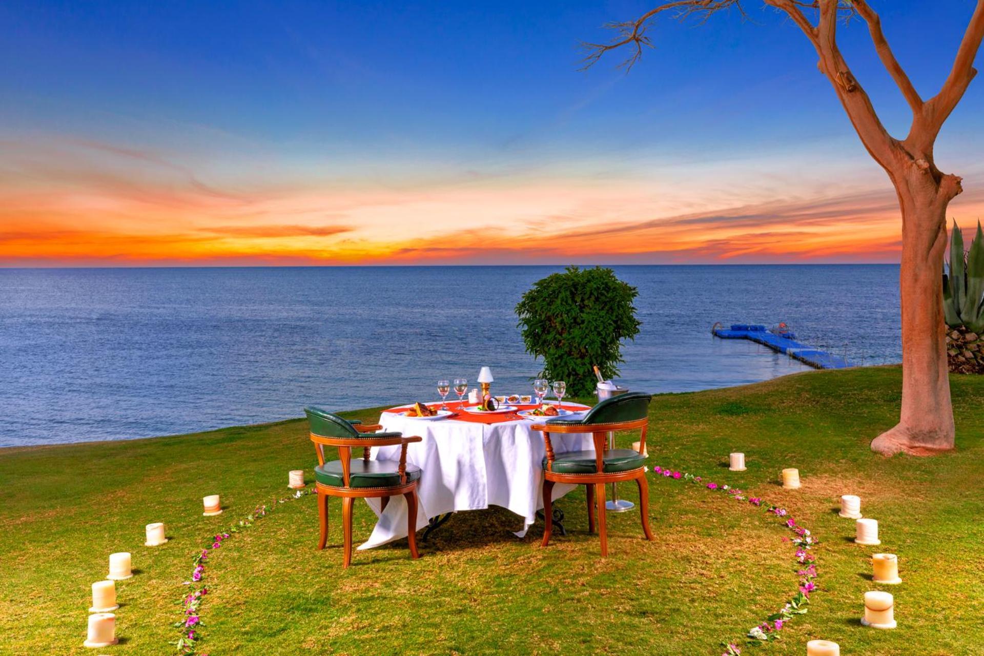 Pickalbatros Royal Grand Sharm - Adults Friendly 16 Years Plus Екстериор снимка The photo depicts a romantic outdoor dining setup by the seaside. In the foreground, there is a round table elegantly set with a white tablecloth, plates, and glassware, creating a cozy atmosphere. Surrounding the table are green chairs. A circle of 