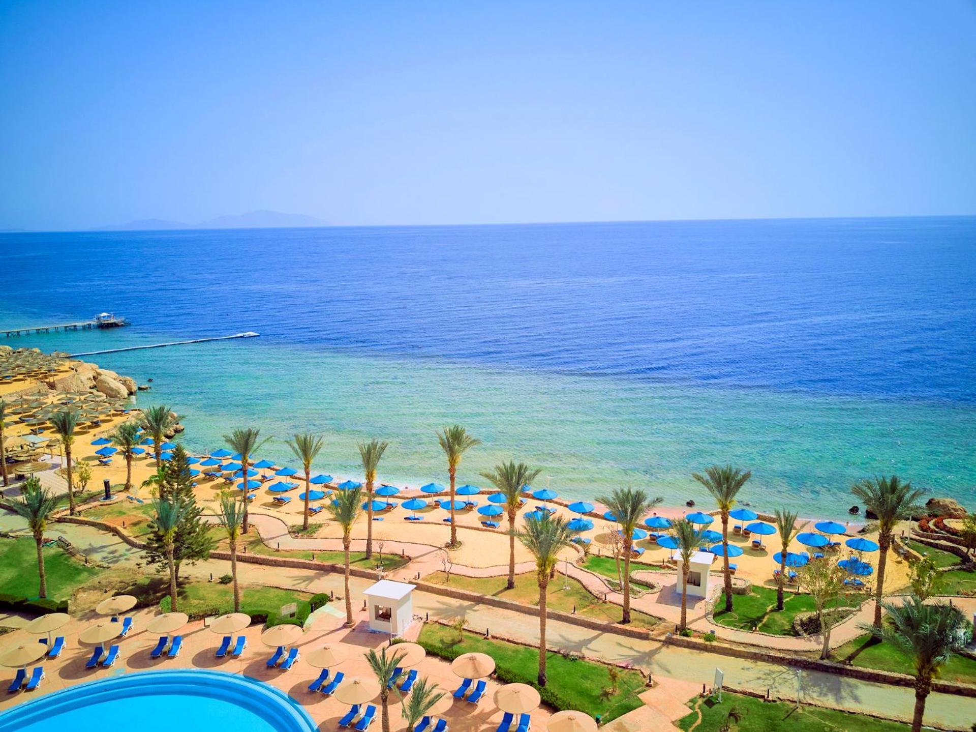 Pickalbatros Royal Grand Sharm - Adults Friendly 16 Years Plus Екстериор снимка The photo displays a beautiful coastal scene featuring a clear blue sea that transitions into green tones near the shore. In the foreground, there are sandy beaches lined with palm trees and a variety of beach umbrellas. There is also a swimming pool