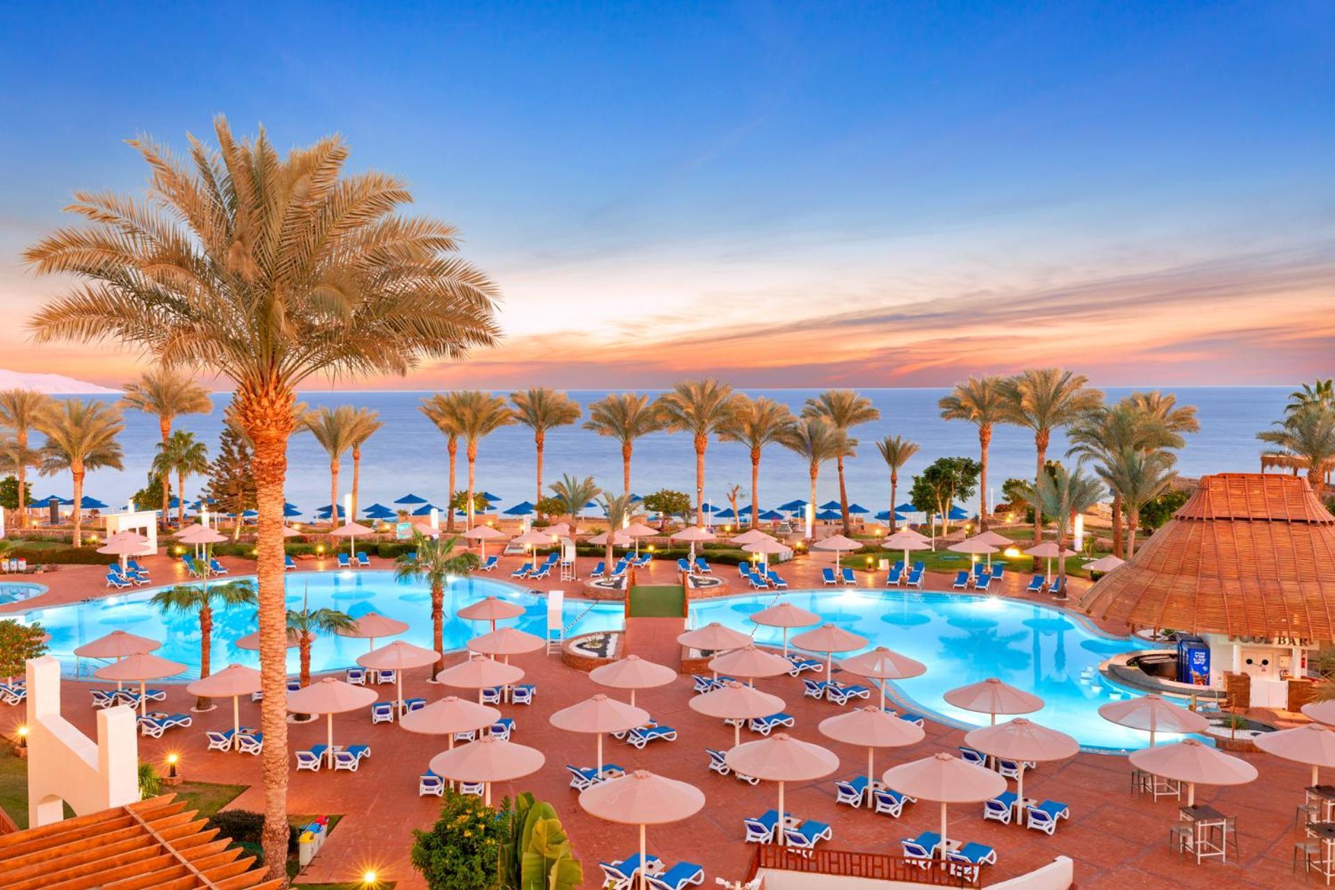 Pickalbatros Royal Grand Sharm - Adults Friendly 16 Years Plus Екстериор снимка The image shows a beautiful resort setting by the beach. There is a large swimming pool at the center, surrounded by lounge chairs and umbrellas. Palm trees line the area, giving it a tropical vibe. In the background, you can see the ocean and a colo