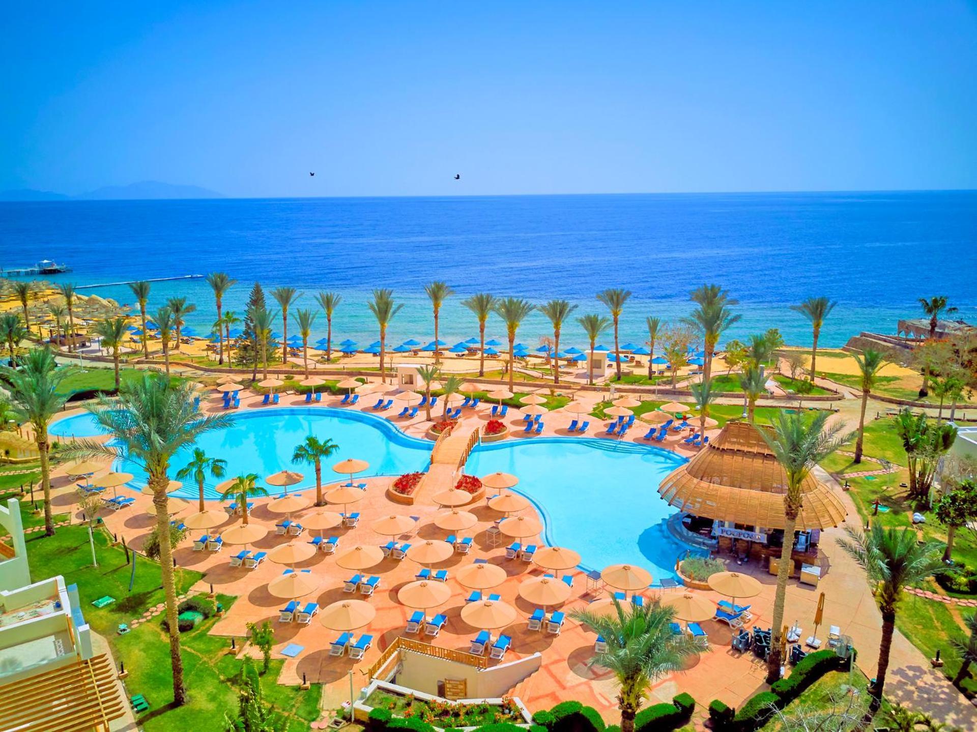 Pickalbatros Royal Grand Sharm - Adults Friendly 16 Years Plus Екстериор снимка The photo depicts a picturesque resort scene by the sea. In the foreground, there is a large, inviting swimming pool surrounded by lounge chairs and umbrellas. Palm trees are strategically placed around the pool area, adding to the tropical atmospher