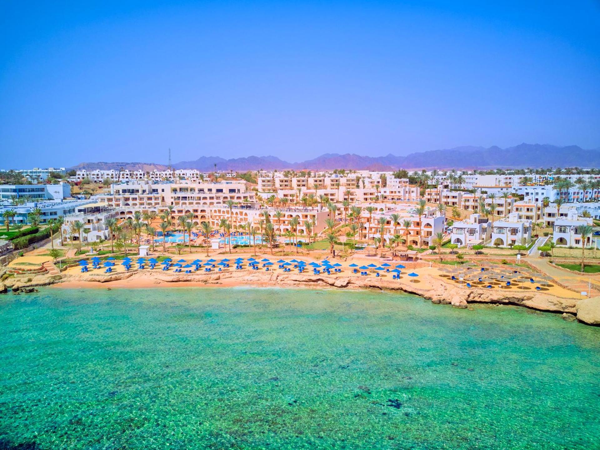 Pickalbatros Royal Grand Sharm - Adults Friendly 16 Years Plus Екстериор снимка The photo depicts a picturesque beachfront resort. It shows a clear turquoise sea with a sandy shore lined with beach umbrellas and sun loungers. Behind the beach, there are several white buildings of the resort, surrounded by lush greenery. In the b