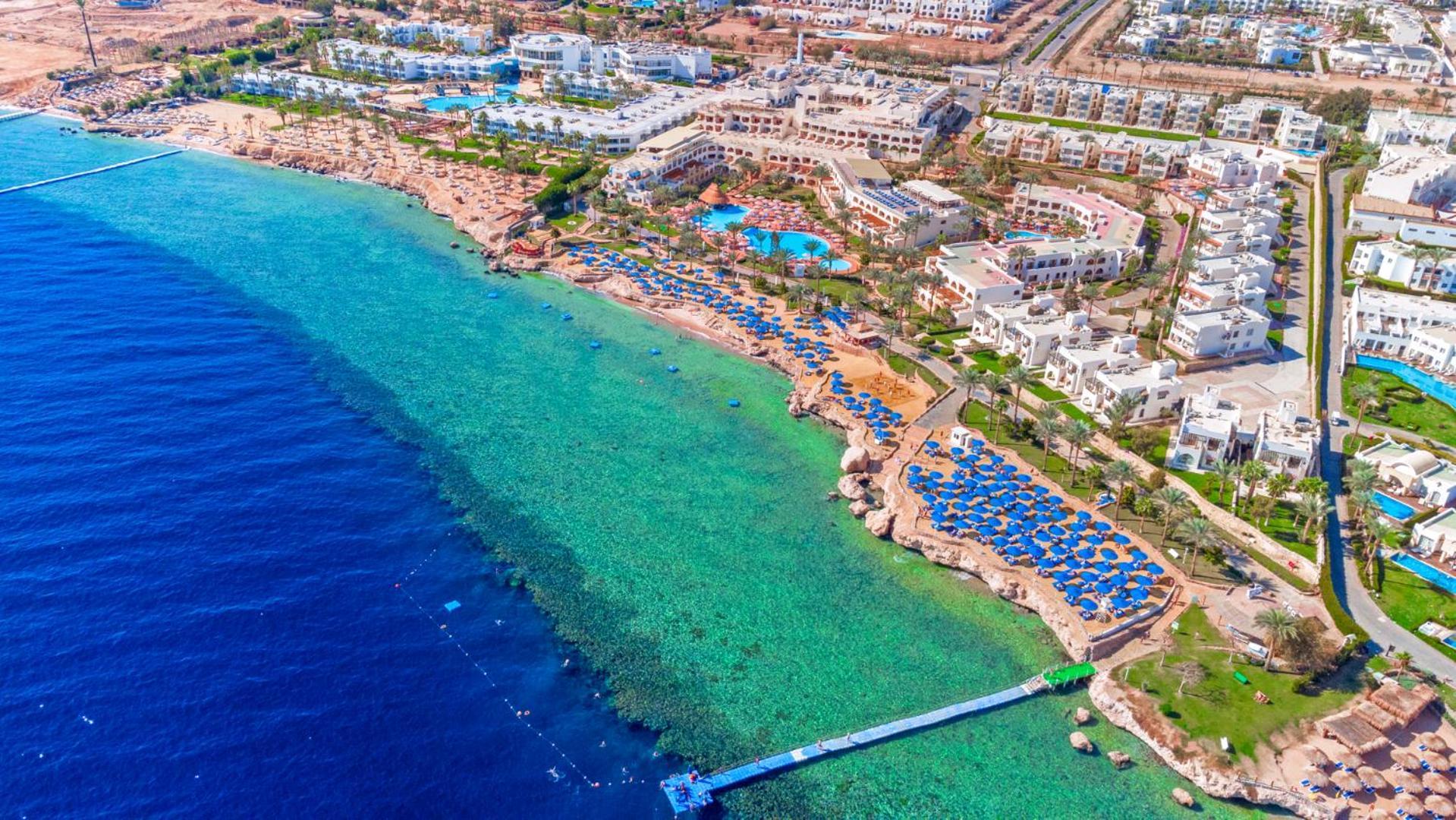 Pickalbatros Royal Grand Sharm - Adults Friendly 16 Years Plus Екстериор снимка The photo showcases a beautiful aerial view of a coastal resort. In the foreground, there's a sandy beach with beach umbrellas and lounge chairs arranged neatly, indicating a tourist area. The clear, turquoise water transitions from the sandy shore t