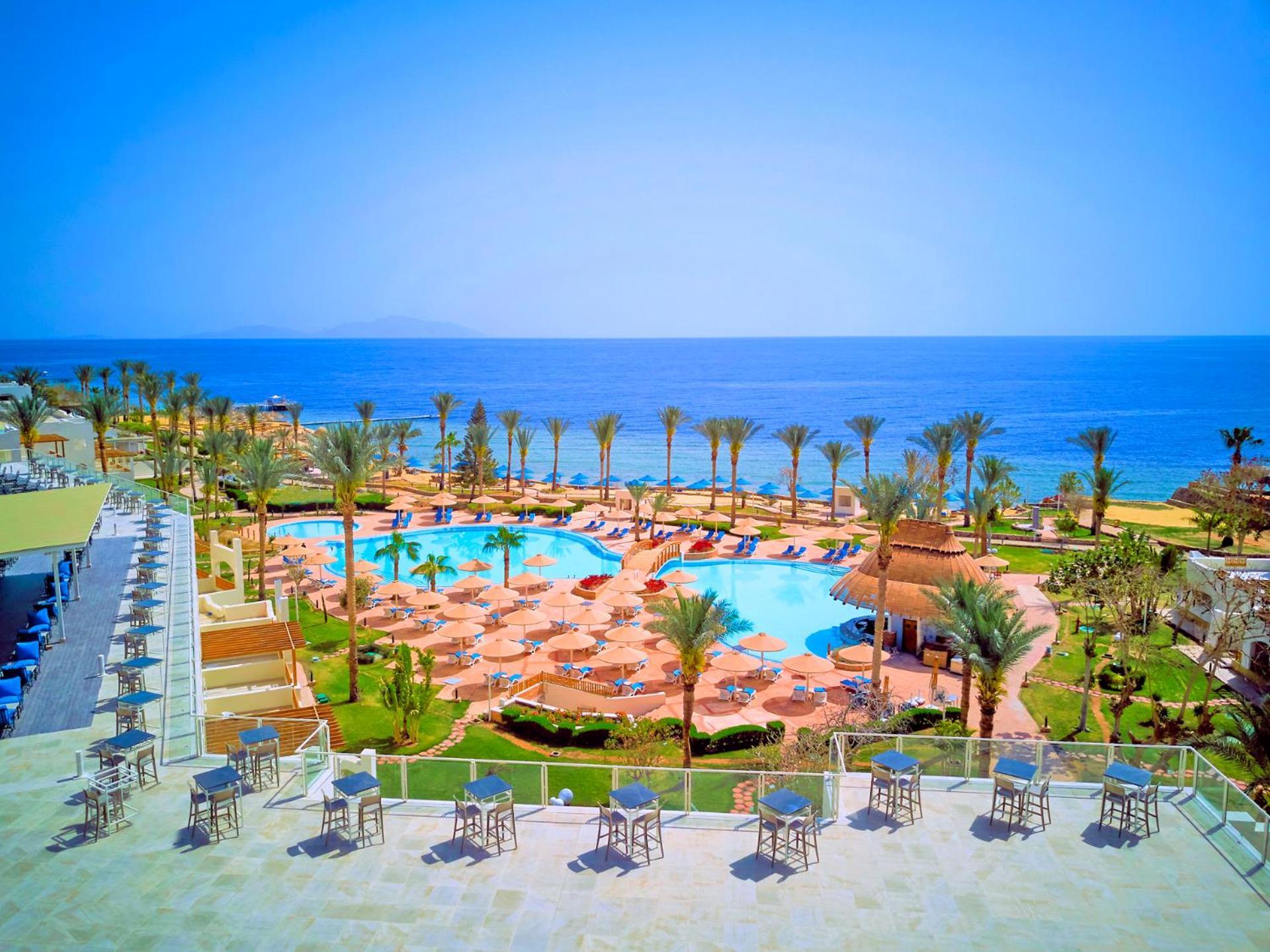 Pickalbatros Royal Grand Sharm - Adults Friendly 16 Years Plus Екстериор снимка The photo shows a beautiful resort scene with a spacious swimming pool at the center, surrounded by sun loungers and palm trees. There are areas for relaxation with tables and chairs positioned around the pool. In the background, the vibrant blue oce