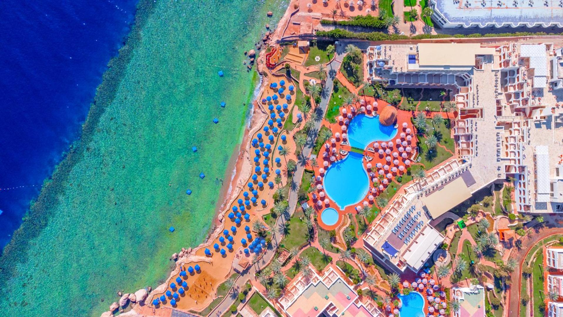 Pickalbatros Royal Grand Sharm - Adults Friendly 16 Years Plus Екстериор снимка The photo showcases an aerial view of a resort area by the sea. It features a turquoise body of water alongside a sandy beach. There are several umbrellas set up on the beach and lounging areas. A large pool is visible, characterized by its unique sh