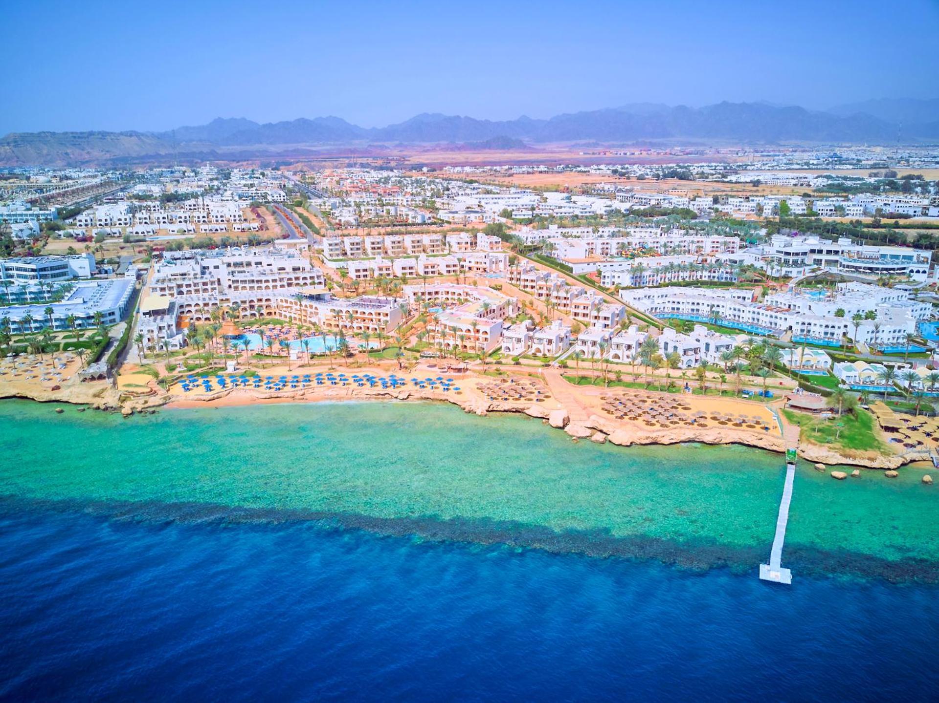 Pickalbatros Royal Grand Sharm - Adults Friendly 16 Years Plus Екстериор снимка The photo shows an aerial view of a coastal resort area. In the foreground, there is a clear blue ocean with a gradient of colors from deep blue to lighter shades near the shore. The beach features sandy areas lined with sun loungers and umbrellas. F