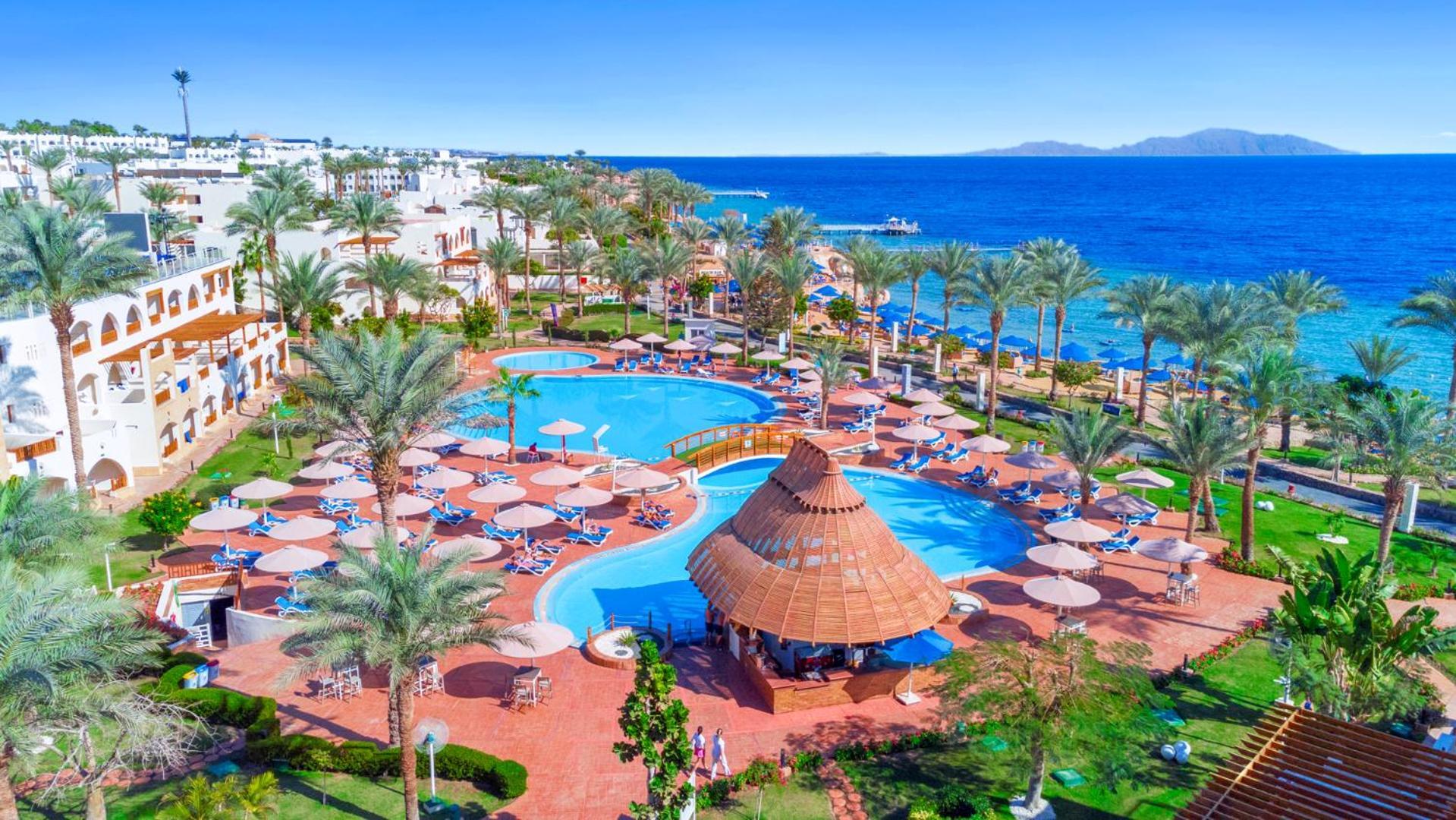 Pickalbatros Royal Grand Sharm - Adults Friendly 16 Years Plus Екстериор снимка The image depicts a vibrant resort scene overlooking a clear blue sea. In the foreground, there are multiple swimming pools surrounded by sun loungers and umbrellas. A thatched-roof gazebo is situated near the center of the pool area. Palms and lush 