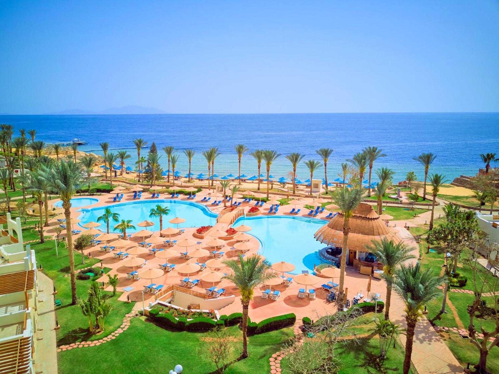 Pickalbatros Royal Grand Sharm - Adults Friendly 16 Years Plus Екстериор снимка The photo depicts a beautiful resort scene by the ocean. In the foreground, there is a large swimming pool surrounded by palm trees and lounge chairs. The pool area features sunbeds and umbrellas, suggesting a relaxing atmosphere for guests. In the b