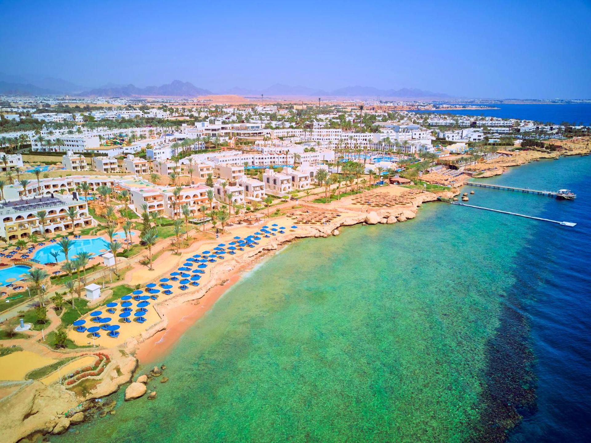 Pickalbatros Royal Grand Sharm - Adults Friendly 16 Years Plus Екстериор снимка The photo showcases a picturesque coastal scene, likely from a resort area. It features a sandy beach lined with blue umbrellas and lounge chairs, indicating a recreational space for relaxation. The water is clear and exhibits shades of green and blu
