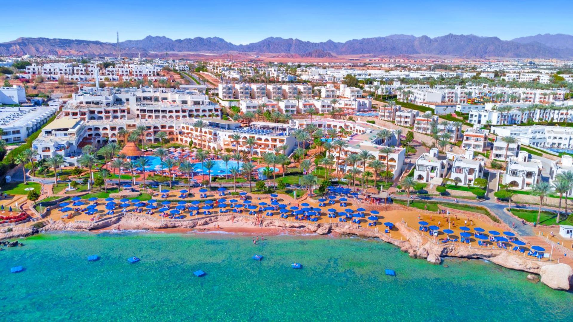 Pickalbatros Royal Grand Sharm - Adults Friendly 16 Years Plus Екстериор снимка The photo showcases a beautiful coastal resort area. In the foreground, there is a sandy beach lined with blue umbrellas and sun loungers. The vibrant turquoise water gently laps at the shore. Behind the beach, you can see a well-maintained resort wi