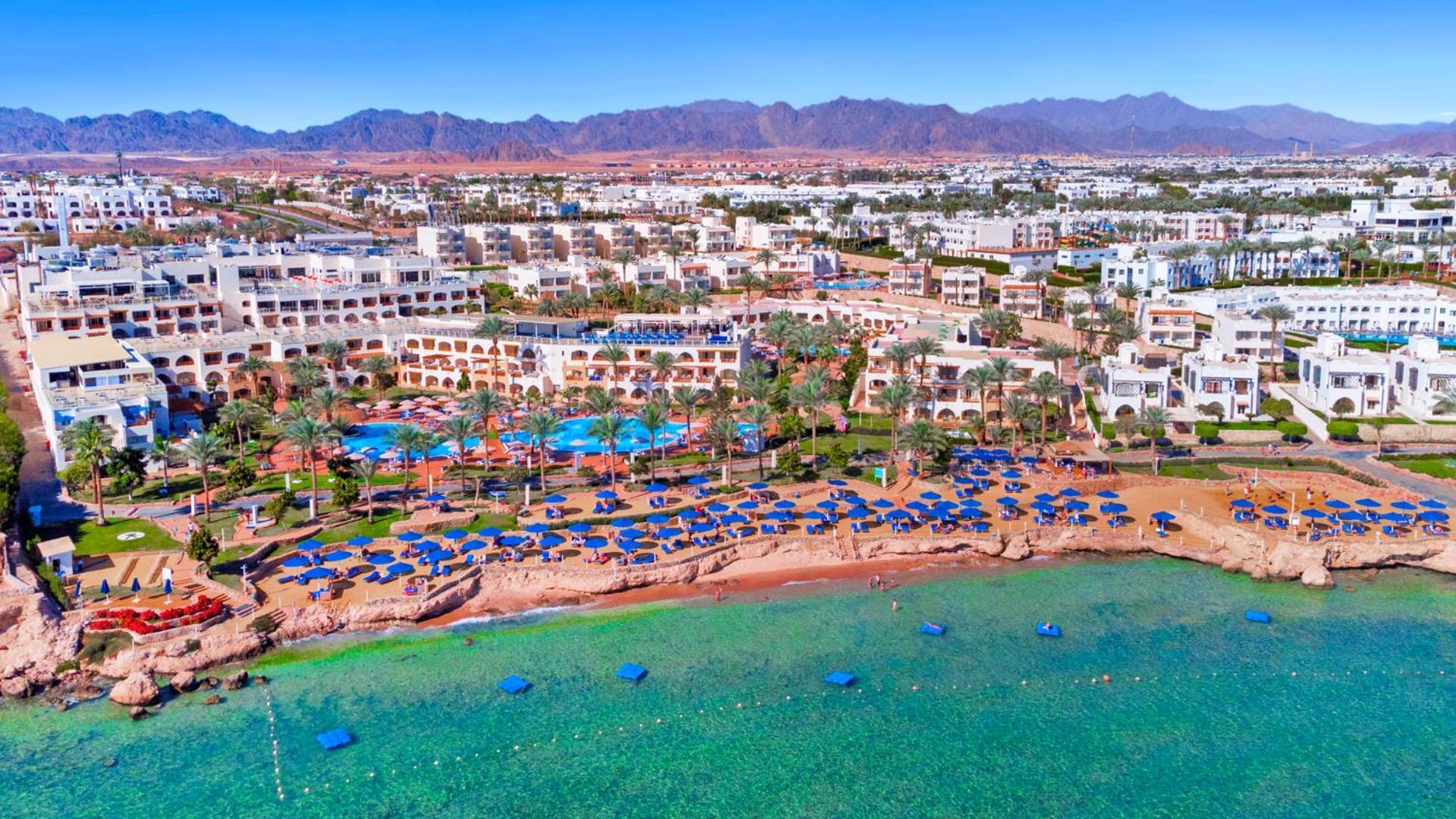 Pickalbatros Royal Grand Sharm - Adults Friendly 16 Years Plus Екстериор снимка The photo shows an aerial view of a coastal resort area. In the foreground, there is a beach with greenish-blue water, where several blue beach umbrellas and lounges are set up for guests. The shoreline appears sandy, and there are some rocky areas. 