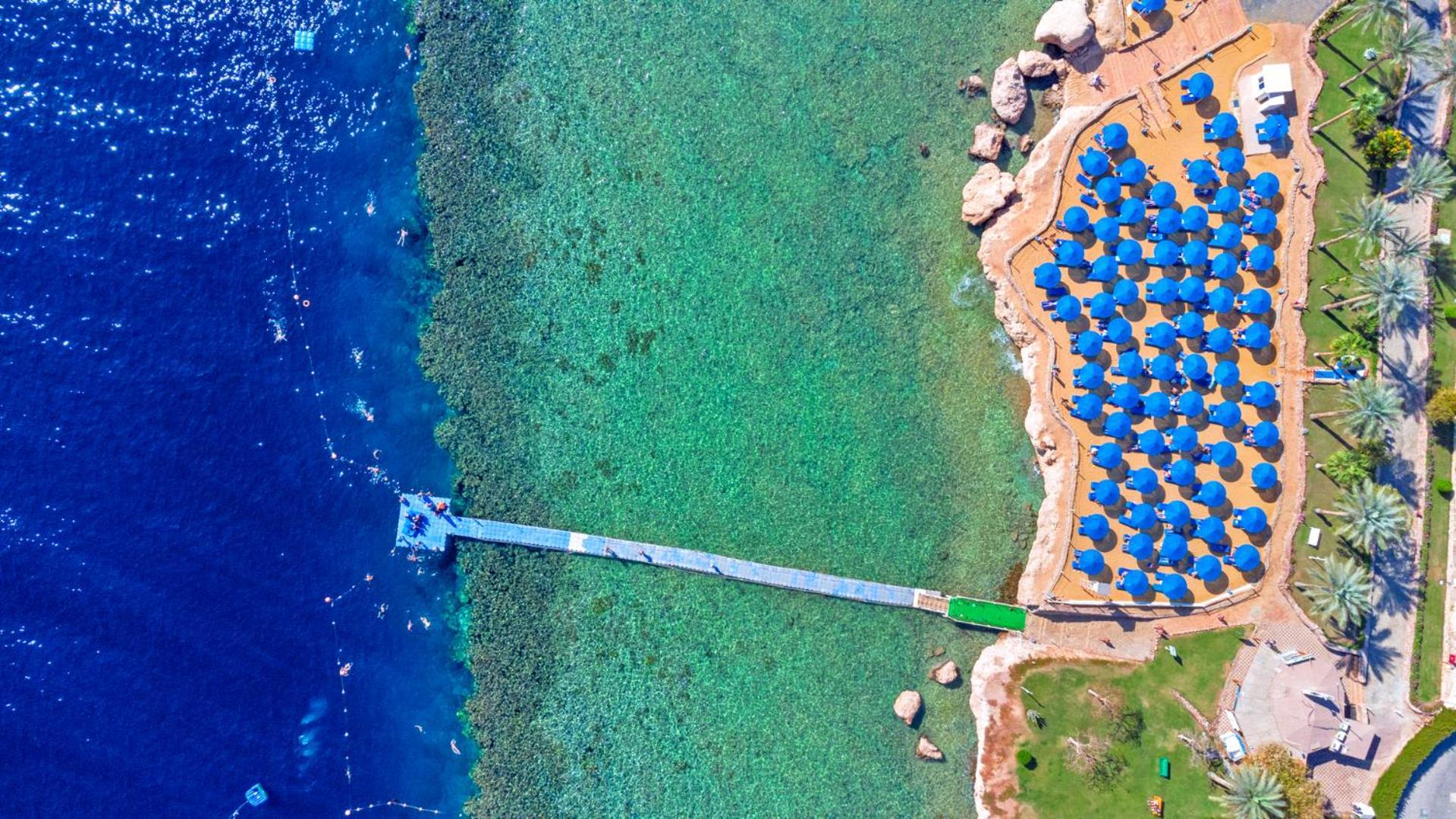 Pickalbatros Royal Grand Sharm - Adults Friendly 16 Years Plus Екстериор снимка The photo shows an aerial view of a beach area. In the foreground, there is a wooden pier extending into the clear blue water. To the right, there are numerous beach umbrellas with blue canopies arranged neatly, likely indicating a resort or beach cl
