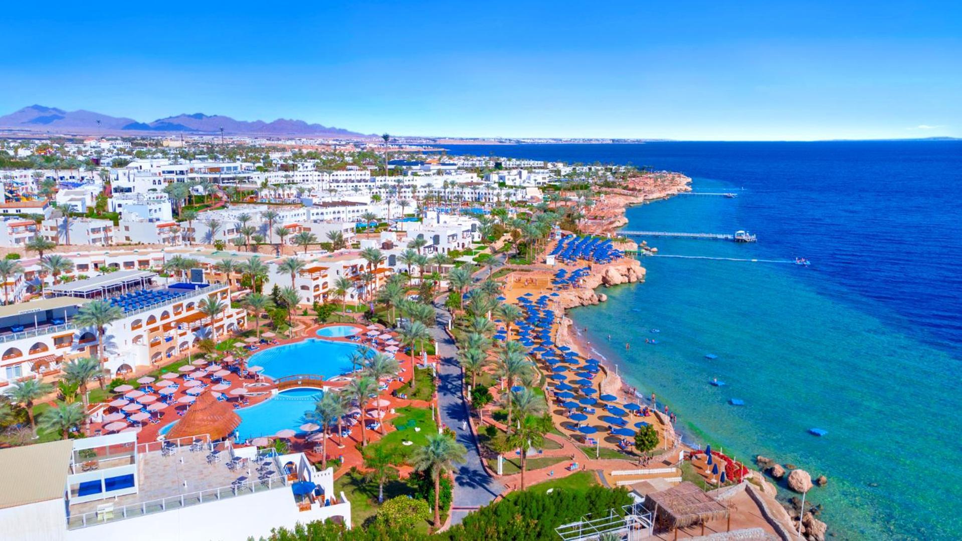 Pickalbatros Royal Grand Sharm - Adults Friendly 16 Years Plus Екстериор снимка The photo features a picturesque coastal scene, likely from a resort area. In the foreground, there is a large swimming pool surrounded by lounge chairs. Further back, the landscape includes several buildings, possibly hotels or apartments, that over
