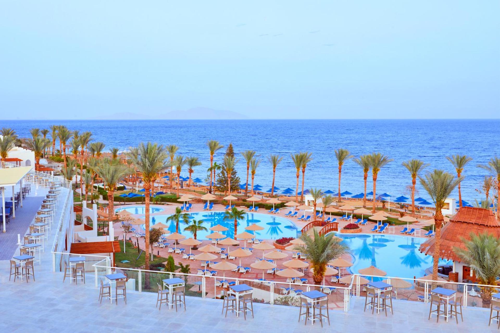 Pickalbatros Royal Grand Sharm - Adults Friendly 16 Years Plus Екстериор снимка The photo depicts a scenic resort area by the sea. In the foreground, there is a large swimming pool surrounded by lounge chairs and umbrellas. Palm trees line the sides of the pool, adding to the tropical atmosphere. In the background, a beautiful b