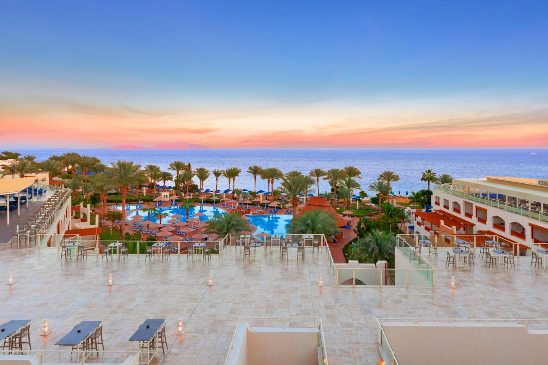 Pickalbatros Royal Grand Sharm - Adults Friendly 16 Years Plus Екстериор снимка The photo depicts a beautiful resort scene at sunset. In the foreground, there is a spacious terrace area with modern furniture. Below, a large swimming pool is surrounded by lush palm trees and lounge chairs, indicating a relaxing atmosphere. The vi