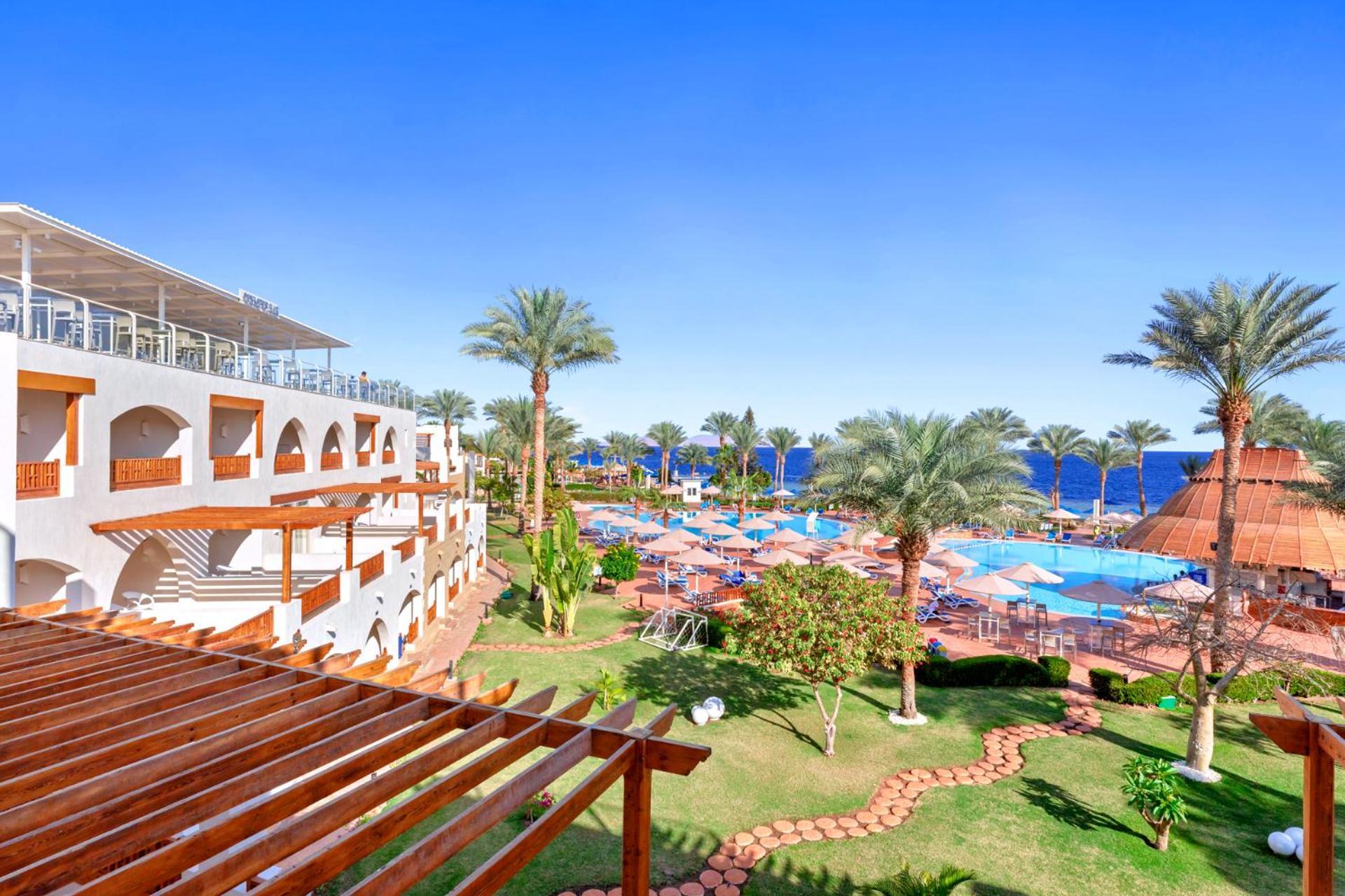 Pickalbatros Royal Grand Sharm - Adults Friendly 16 Years Plus Екстериор снимка The photo showcases a beautiful resort setting, featuring several palm trees and a clear blue sky. In the foreground, there's an area with landscaped gardens, including green grass and a stone pathway. The resort buildings, with archways and wooden d