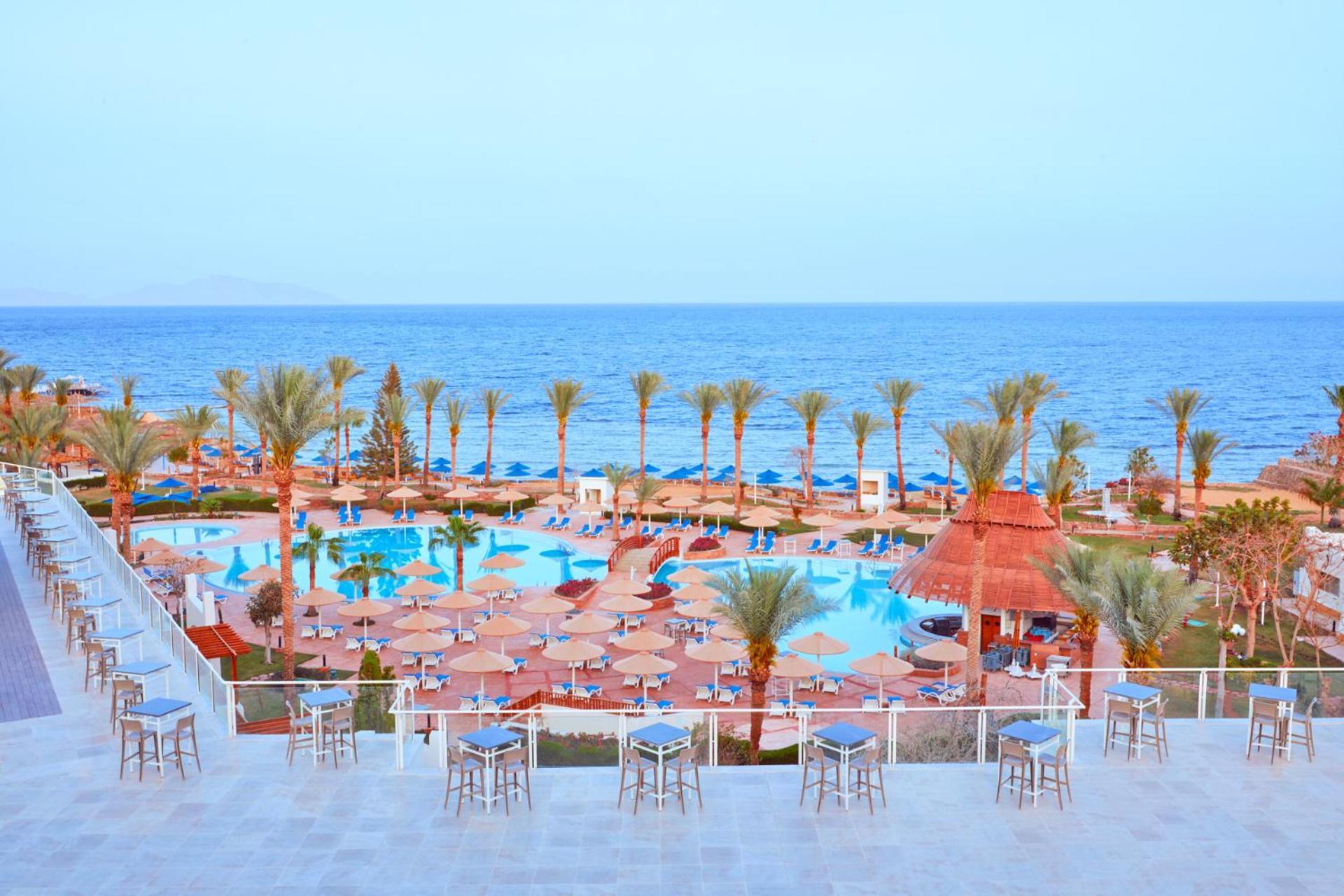 Pickalbatros Royal Grand Sharm - Adults Friendly 16 Years Plus Екстериор снимка The photo shows a scenic view of a resort or beach area. In the foreground, there are several seating areas with bar stools on a terrace. Below, a large swimming pool is visible, surrounded by lounging chairs and umbrellas. Palm trees line the area, 