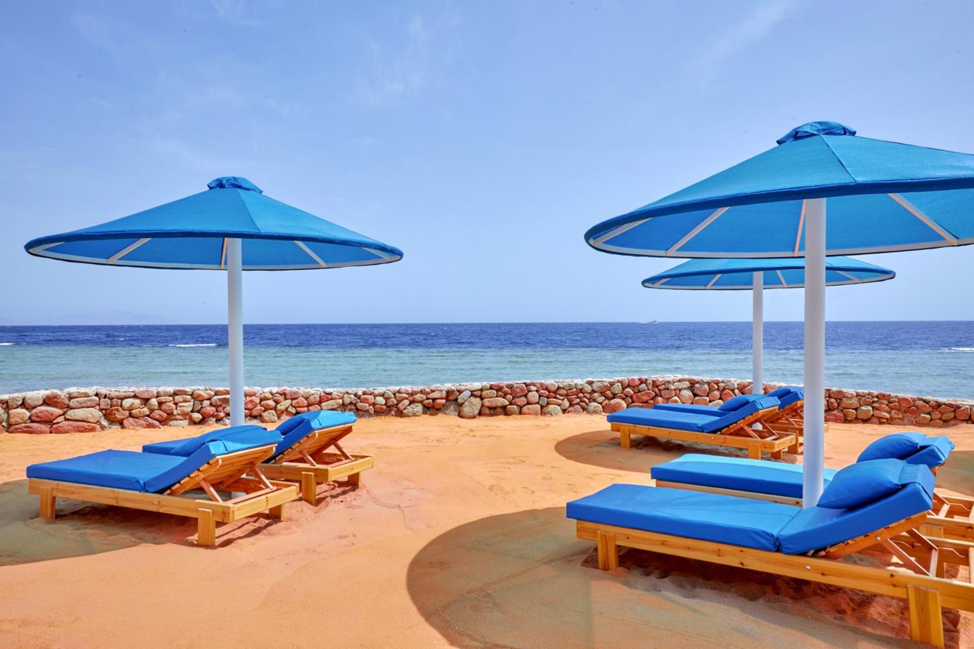 Pickalbatros Royal Grand Sharm - Adults Friendly 16 Years Plus Екстериор снимка The photo shows a beach scene featuring several blue umbrellas and lounge chairs on a sandy area. The umbrellas are large and have a bright blue color, providing shade for the lounge chairs underneath. In the background, there's a view of the ocean w