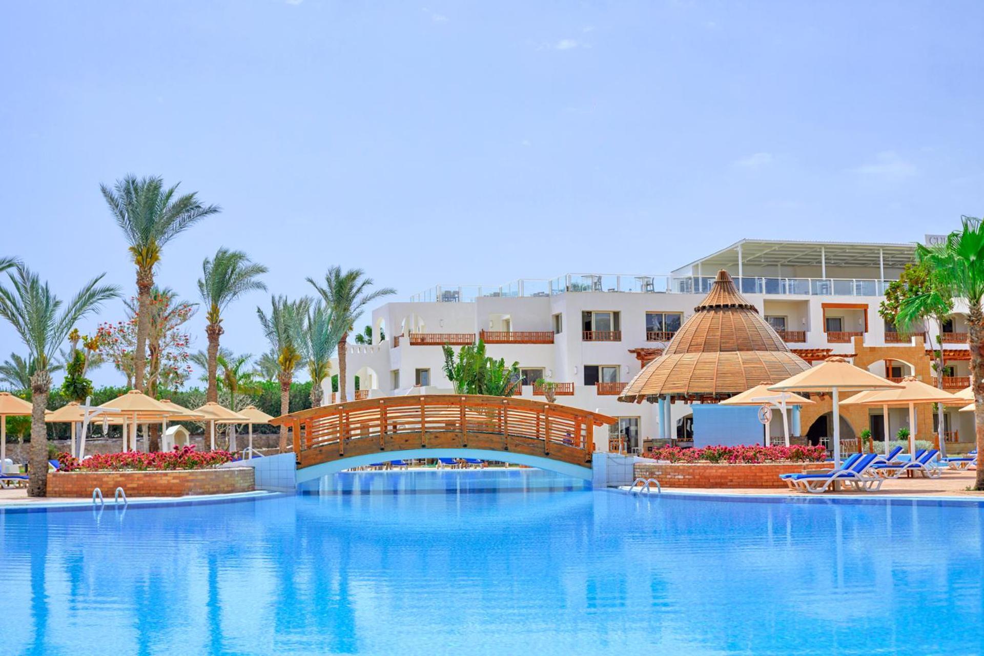 Pickalbatros Royal Grand Sharm - Adults Friendly 16 Years Plus Екстериор снимка The photo depicts a serene resort setting featuring a large, calm swimming pool, which reflects the clear blue sky. In the foreground, a wooden bridge arches over the pool. Surrounding the pool are several palm trees and tropical foliage, contributin