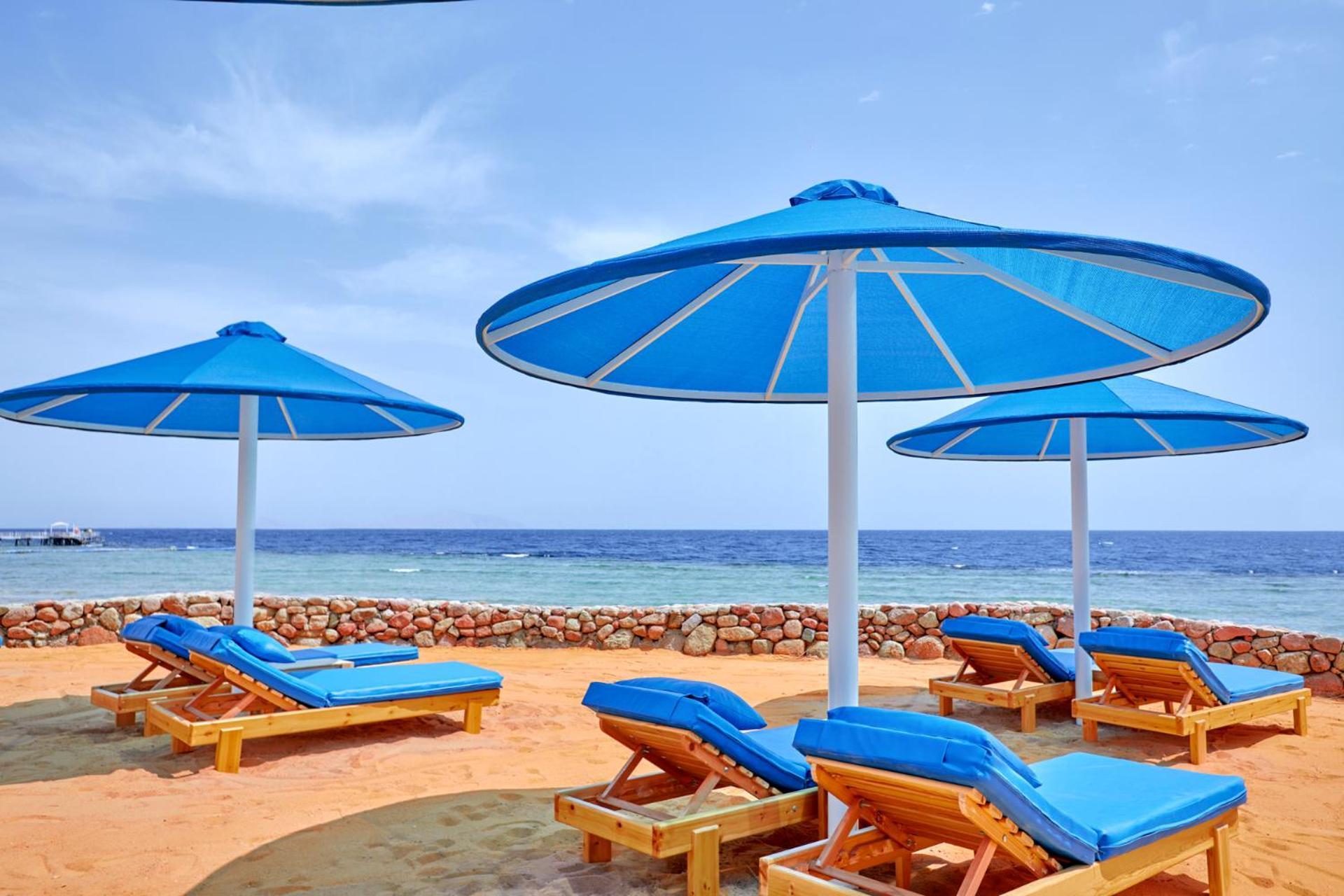 Pickalbatros Royal Grand Sharm - Adults Friendly 16 Years Plus Екстериор снимка The photo shows a serene beach scene with several blue sun loungers arranged beneath large blue umbrellas. The loungers are positioned on sandy ground, with the ocean visible in the background. The sky is clear, indicating a bright and sunny day, and
