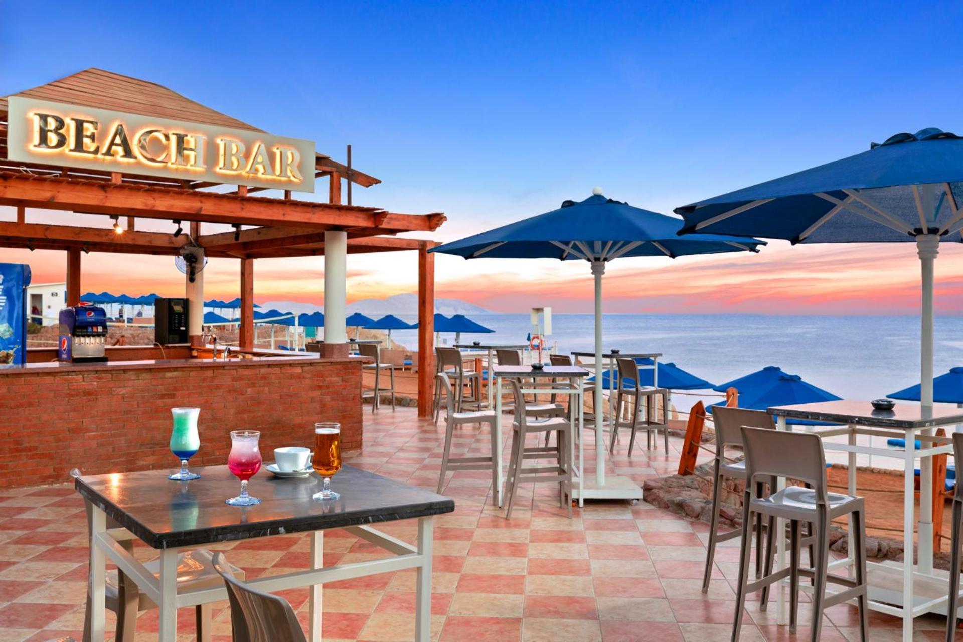 Pickalbatros Royal Grand Sharm - Adults Friendly 16 Years Plus Екстериор снимка The photo shows a beach bar setting, featuring a wooden structure with the words "BEACH BAR" prominently displayed. There are several tables and chairs arranged on a tiled floor, with some drinks placed on the tables. Blue umbrellas provide shade for