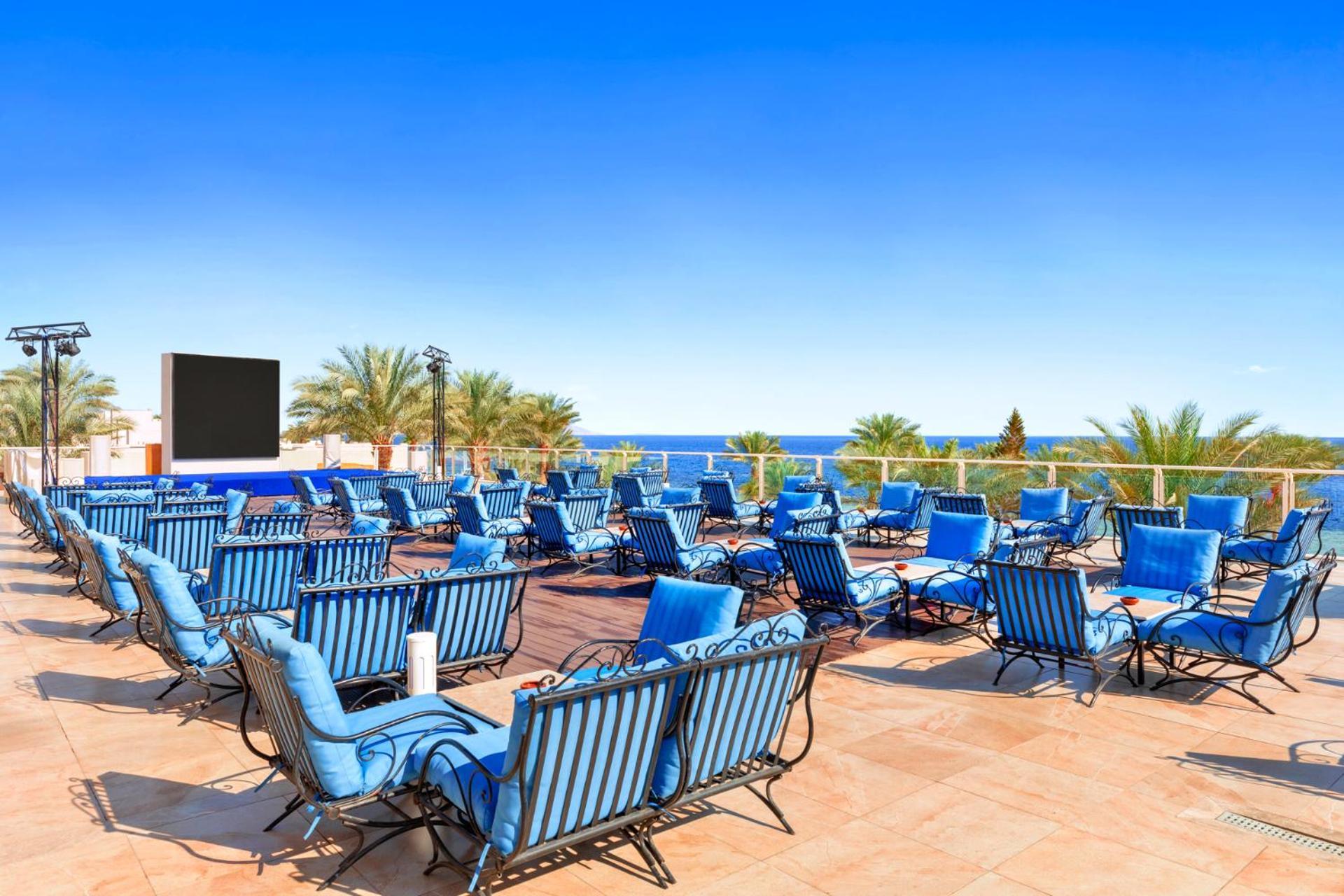 Pickalbatros Royal Grand Sharm - Adults Friendly 16 Years Plus Екстериор снимка The photo features a sunny outdoor space with numerous blue lounge chairs arranged in rows on a tiled patio. In the background, there is a clear blue sky and a view of the ocean, along with some palm trees adding to the tropical atmosphere. Additiona