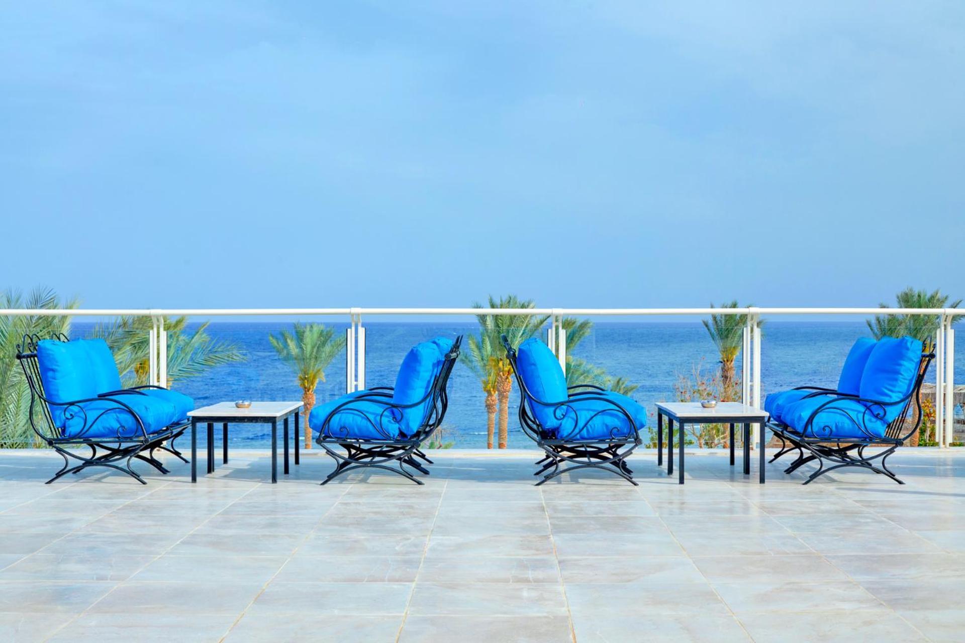 Pickalbatros Royal Grand Sharm - Adults Friendly 16 Years Plus Екстериор снимка The photo depicts a serene outdoor patio area with four blue lounge chairs arranged around small tables. The setting features a clear view of the ocean with palm trees visible in the background, complementing the tranquil atmosphere. The overall colo