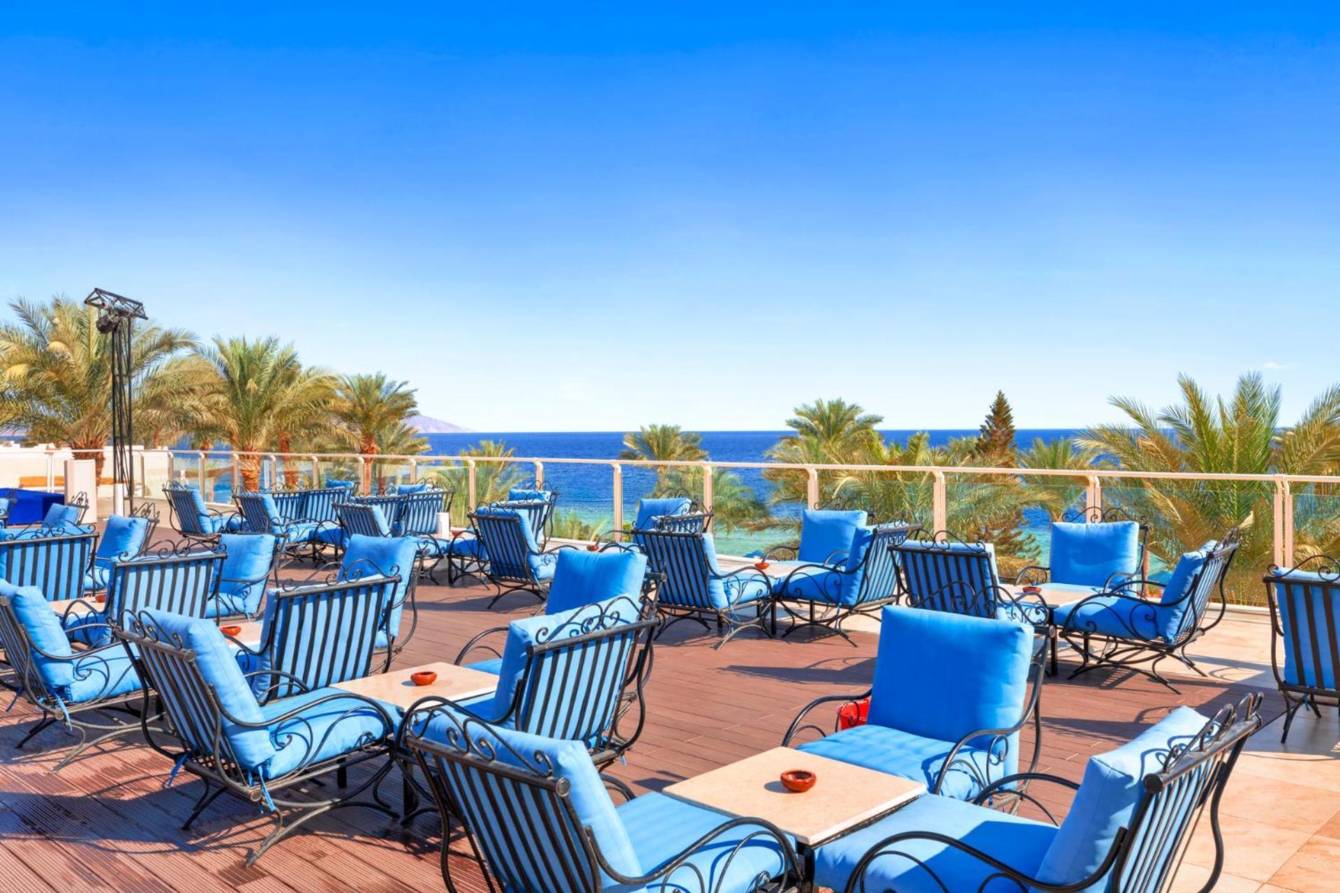 Pickalbatros Royal Grand Sharm - Adults Friendly 16 Years Plus Екстериор снимка The photo shows an outdoor deck or terrace area, adorned with numerous blue lounge chairs arranged neatly. In the background, there are palm trees and a clear blue sea that stretches toward the horizon under a bright sky. The setting appears to be a 