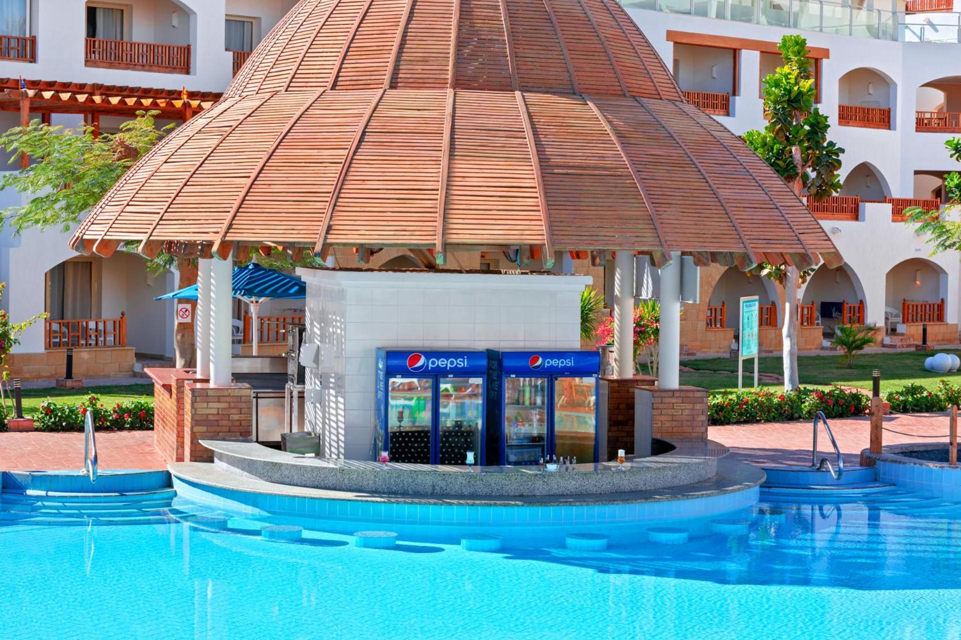Pickalbatros Royal Grand Sharm - Adults Friendly 16 Years Plus Екстериор снимка The photo shows a poolside bar with a thatched roof structure, located near a swimming pool. The bar features a counter where drinks are served, and there are vending machines displaying the Pepsi brand. Surrounding the bar, there are neatly landscap